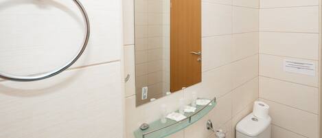 A bathroom with a glass shelf holding toiletries, a toilet, and a sink with a mirror above it.