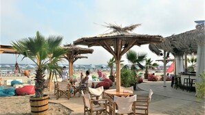 A beachside terrace with wooden tables and chairs, palm trees, and a view of the ocean. - New Line Village Apartments (Sunny Beach)