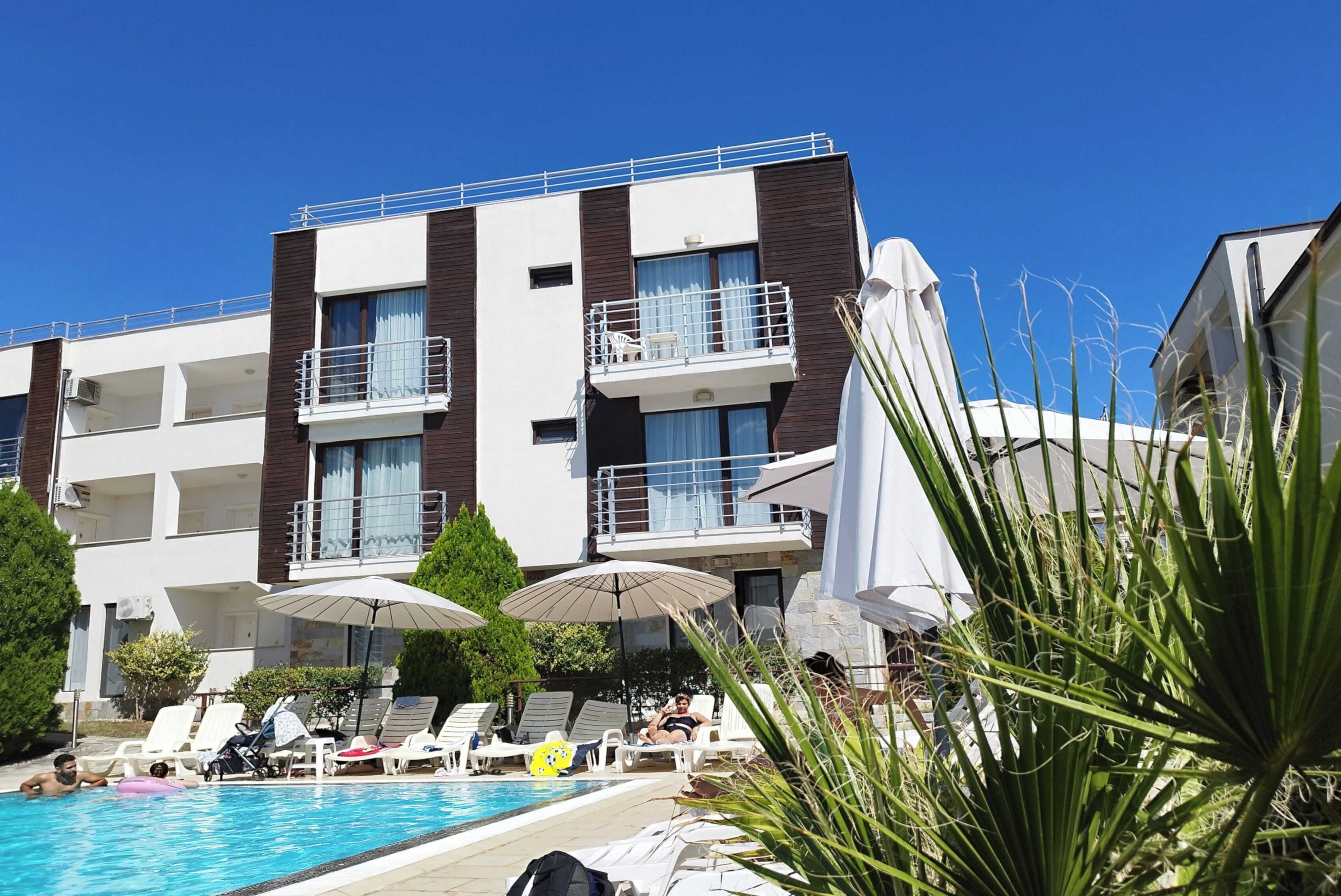 Un hotel moderno con zona de piscina, tumbonas, sombrillas y un cielo azul despejado.