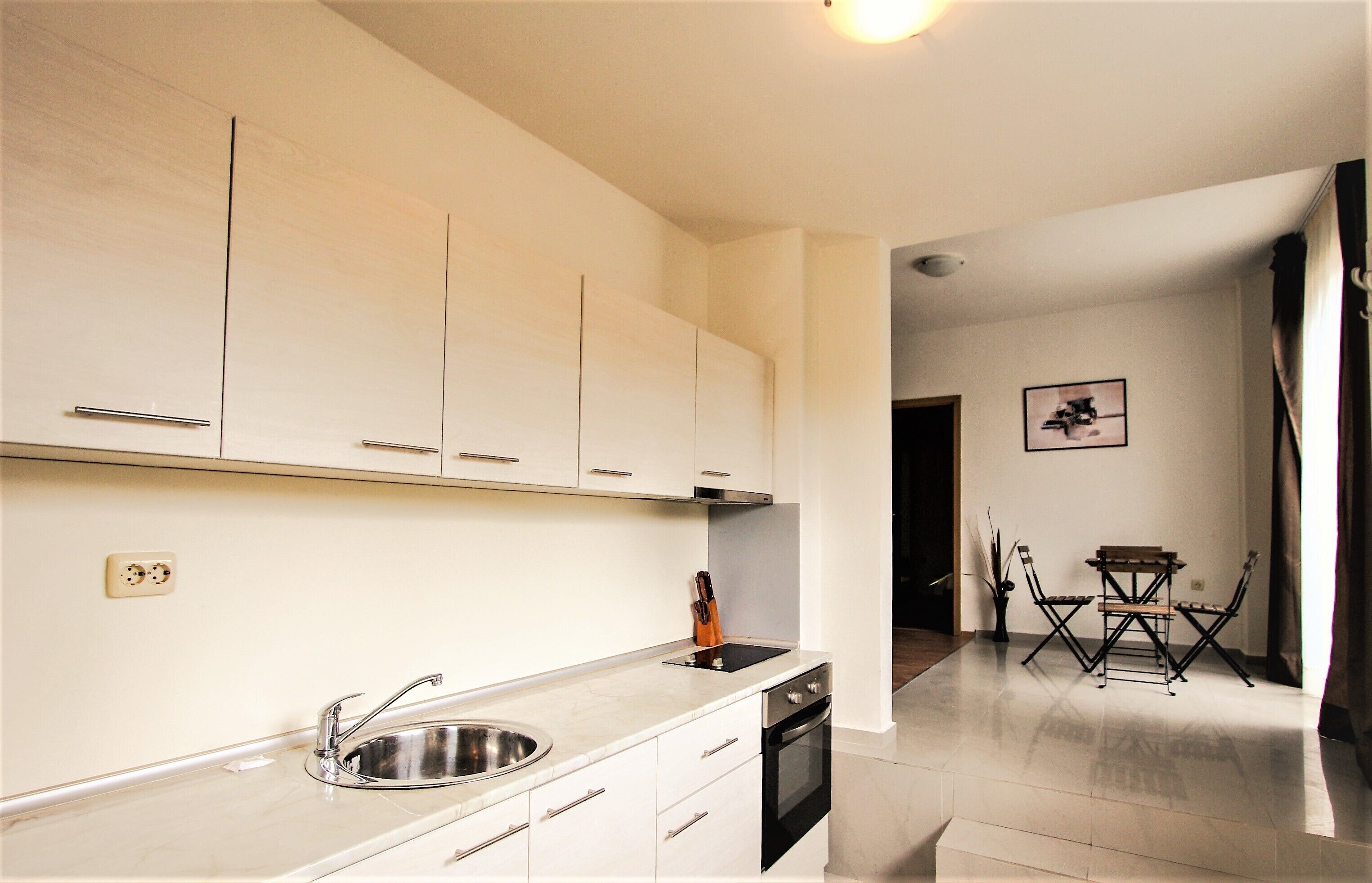 A modern kitchen with white cabinets, a stainless steel sink, and an oven. There is a dining area with a table and chairs.
