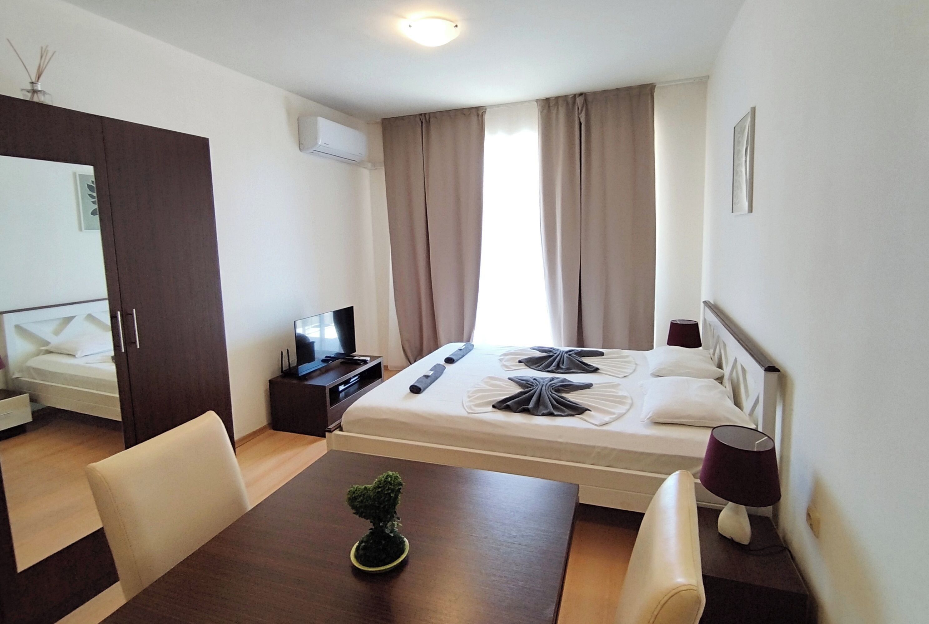 Hotellihuone, jossa on sänky, työpöytä, tuoli, pieni pöytä, jossa on kasvi, televisio ja ikkuna, jossa on verhot.