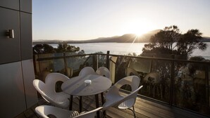 Un balcone con ringhiera in vetro, sedie bianche e un tavolo, con vista su uno specchio d'acqua e colline lontane.