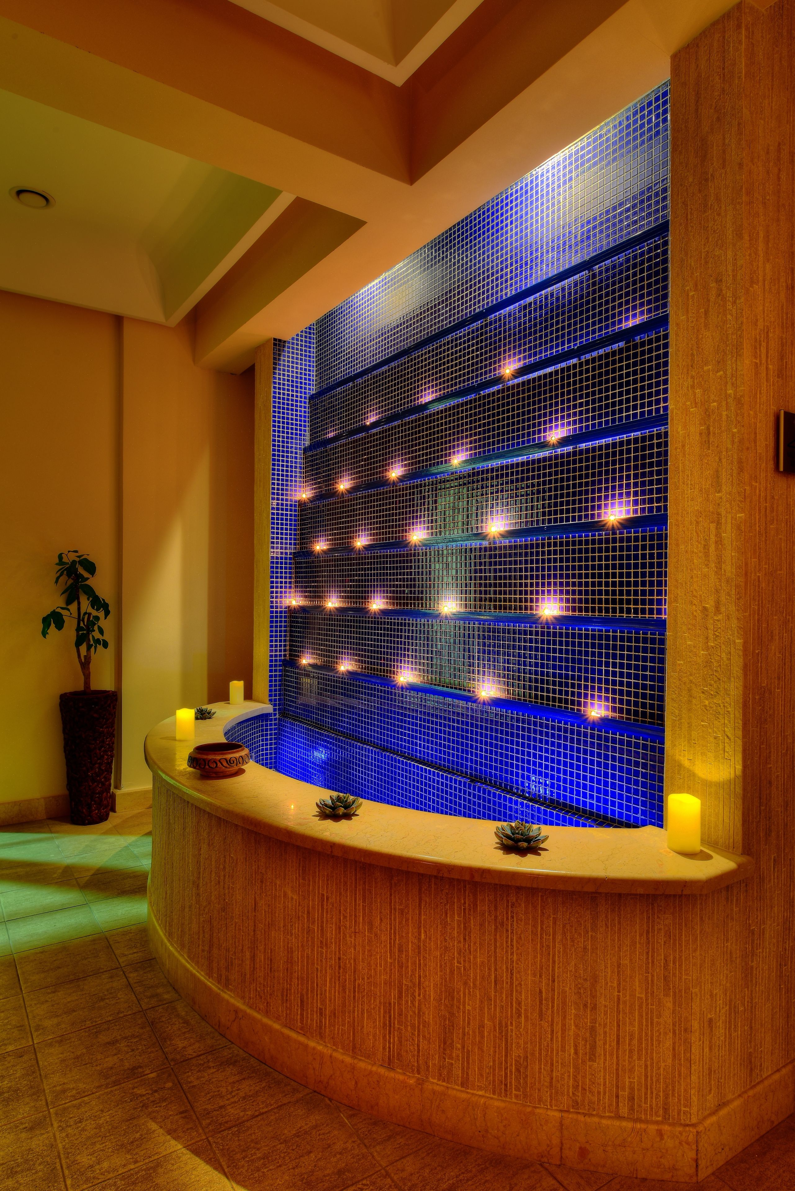 A spa area with a curved reception desk, a wall with blue mosaic tiles, and a potted plant.