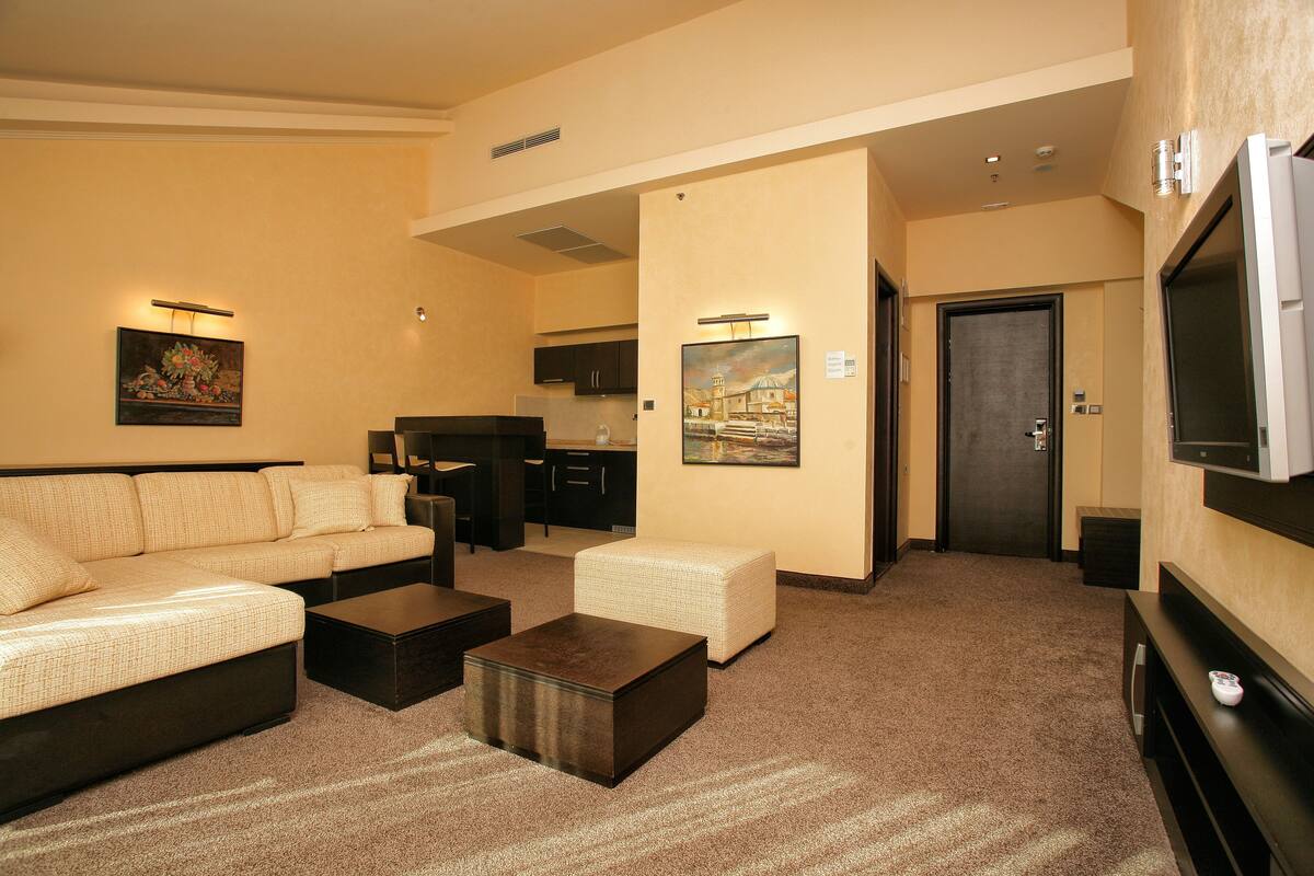 A hotel room with a sofa, a coffee table, a TV, a painting, and a kitchenette area.