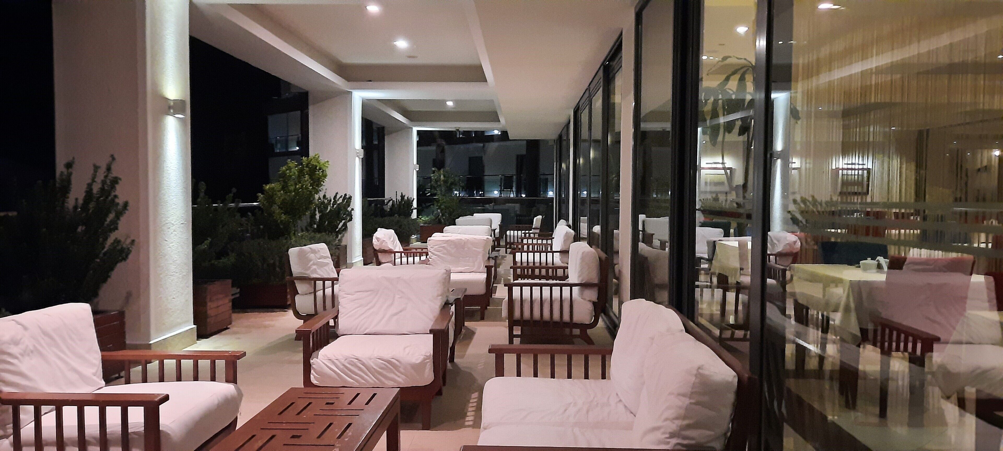 A spacious outdoor seating area with white cushioned chairs and tables, surrounded by glass walls and potted plants.