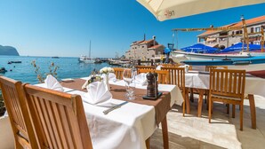 An outdoor dining area with tables set for two, overlooking a marina with boats and a historic building in the background. - Monte Casa Spa & Wellness (Petrovac)