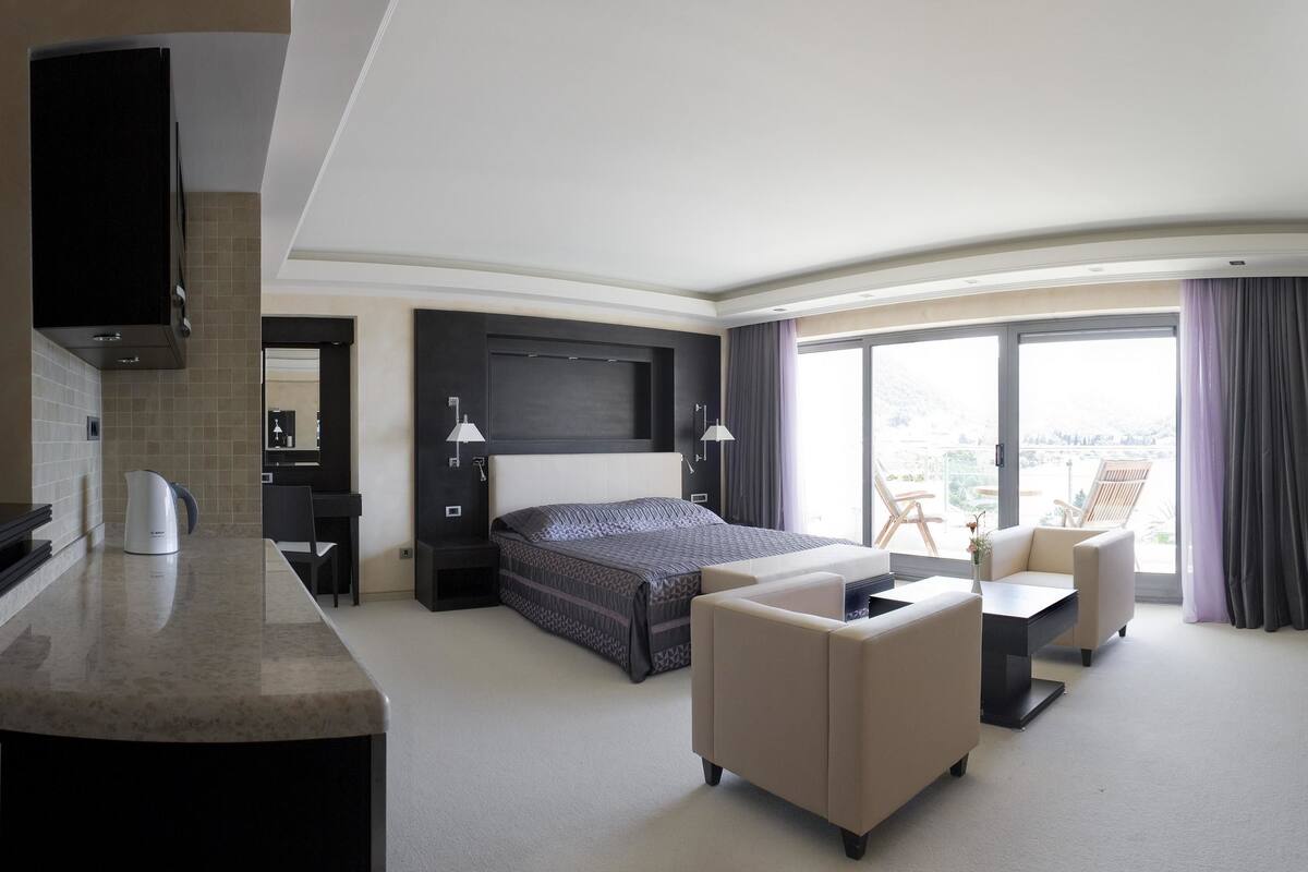 A modern hotel room with a bed, a desk, a chair, a sofa, a TV, and a large window with a view.
