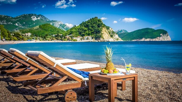 A beach with wooden lounge chairs and a table with fruit and a drink, overlooking a clear blue sea and green hills. - Monte Casa Spa & Wellness (Petrovac)