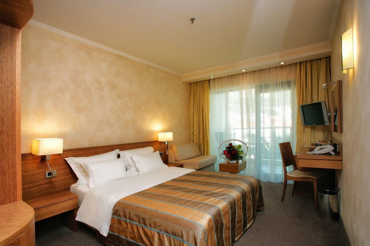 A hotel room with a large bed, a desk with a chair, a television, and a desk lamp.
