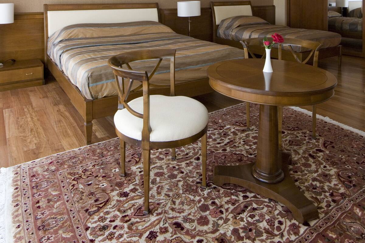 A hotel room with a large bed, a smaller bed, a round table with a chair, a small table with a vase, and a patterned rug.