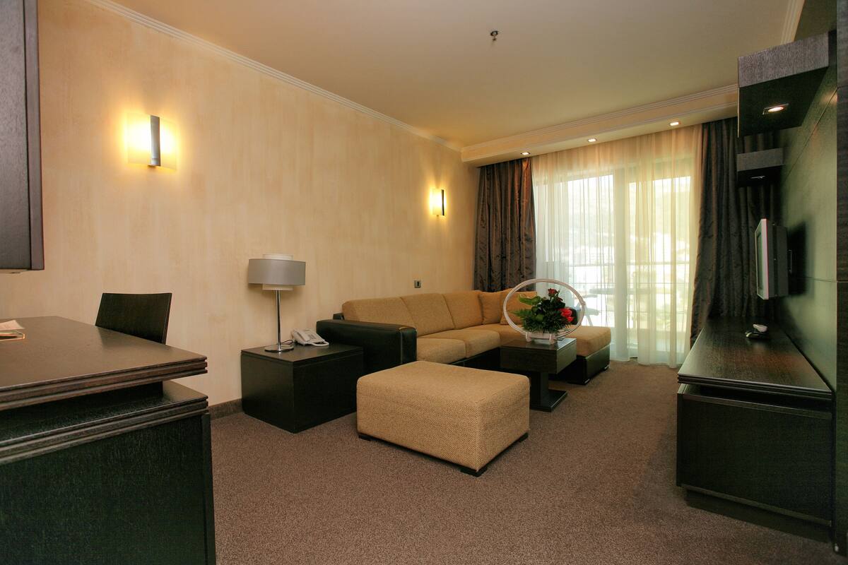 A hotel room with a sofa, ottoman, coffee table, TV, and a desk with a lamp.