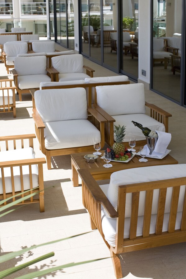 A sunlit outdoor seating area with wooden chairs and tables, a bottle of champagne, and a fruit platter.