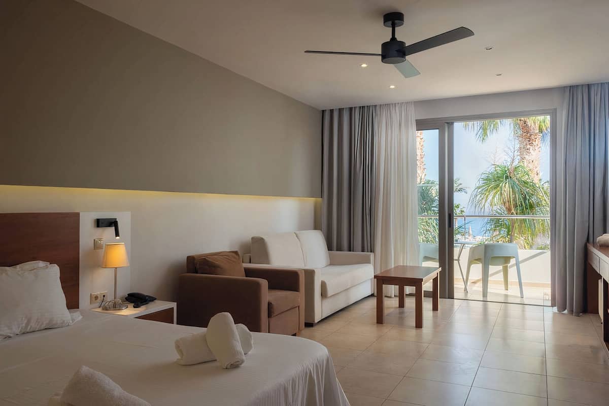 A hotel room with a bed, a sofa, a small table, a nightstand with a lamp, and a balcony with a view of palm trees.