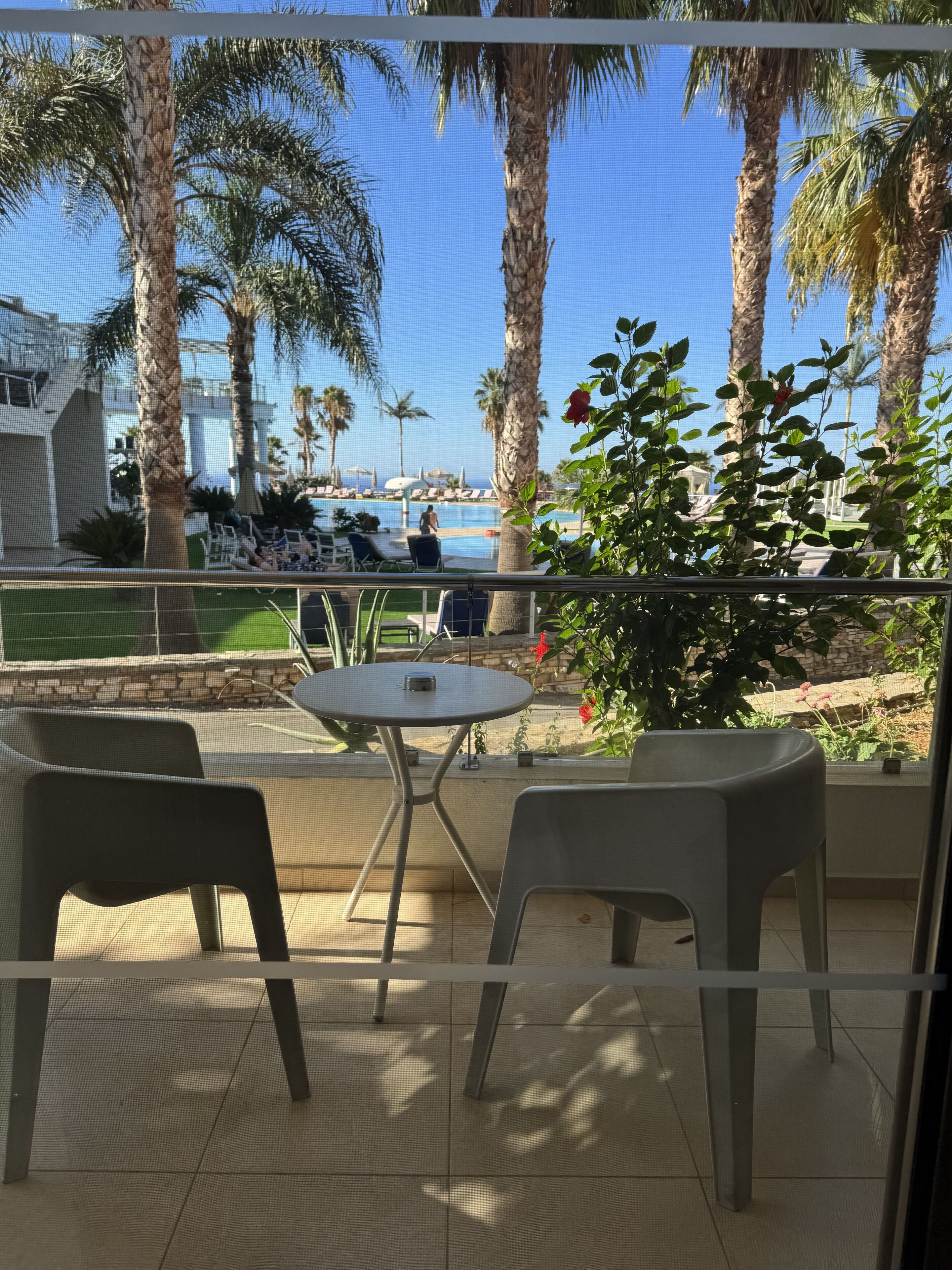 Un balcón con dos sillas y una pequeña mesa, con vista a un área de piscina con palmeras y sillones de descanso.