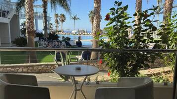 Un balcón con dos sillas y una pequeña mesa, con vista a un área de piscina con palmeras y sillones de descanso.