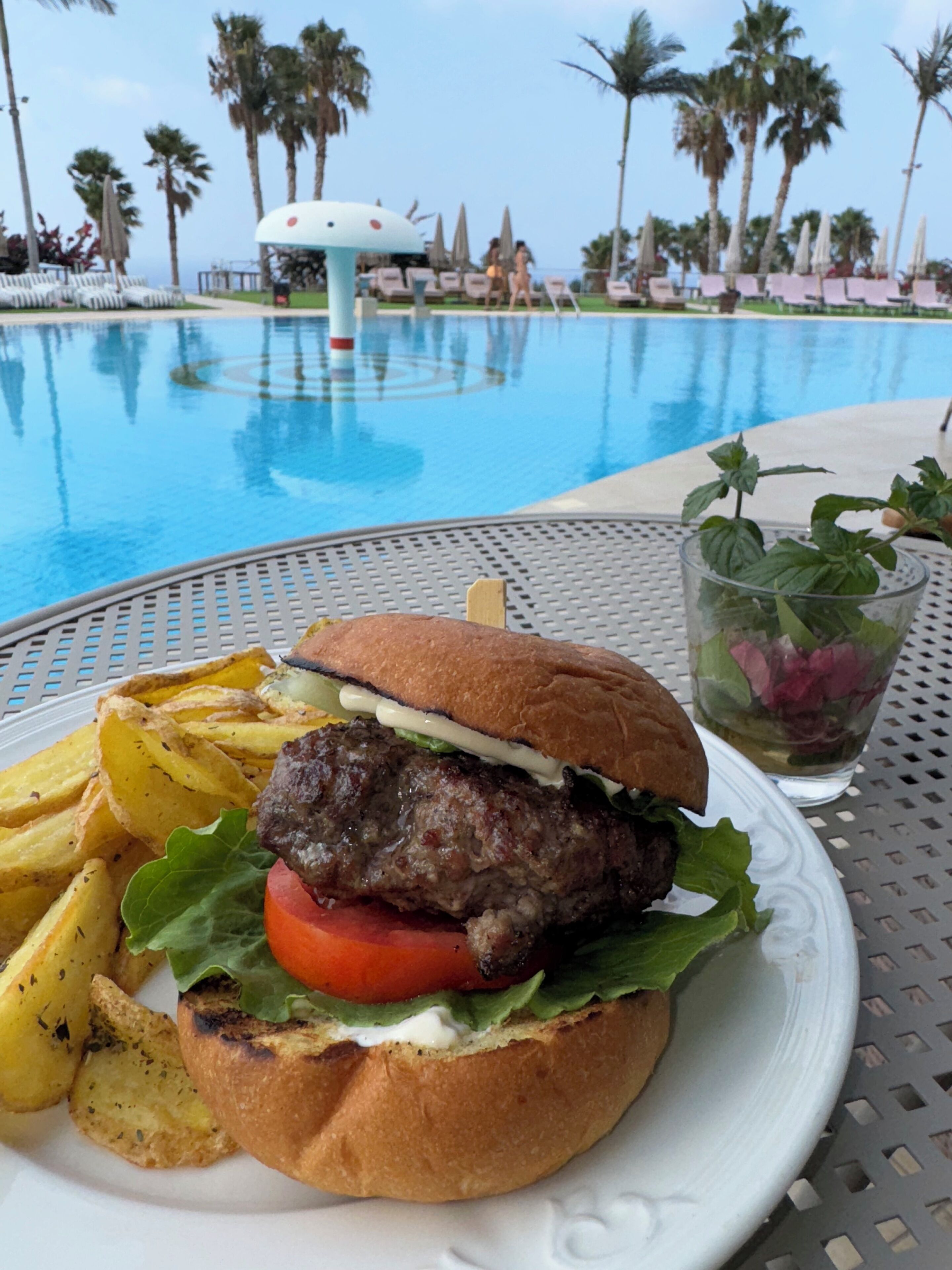 A meal with a burger, fries, and a drink, with a pool and palm trees in the background.
