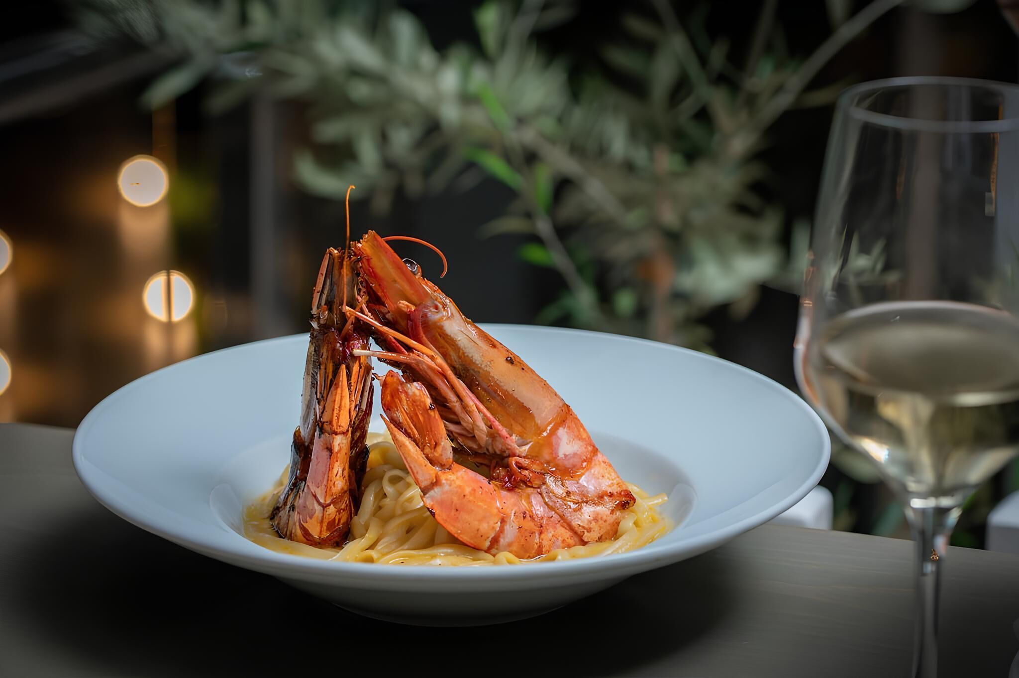 Ein Teller Pasta mit gegrillten Garnelen, ein Glas Weißwein und ein unscharfer Hintergrund mit Grünpflanzen.