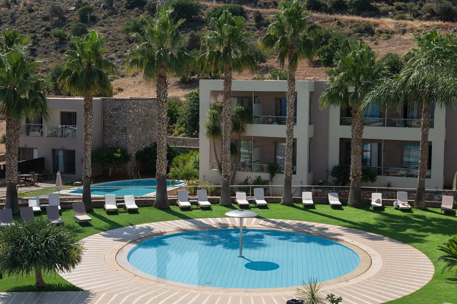 Un hotel moderno con piscina, palmeras y montañas al fondo.