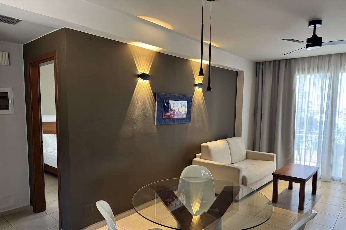 A modern hotel room with a glass dining table, white chairs, a sofa, and a ceiling fan.