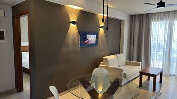 A modern hotel room with a glass dining table, white chairs, a sofa, and a ceiling fan.