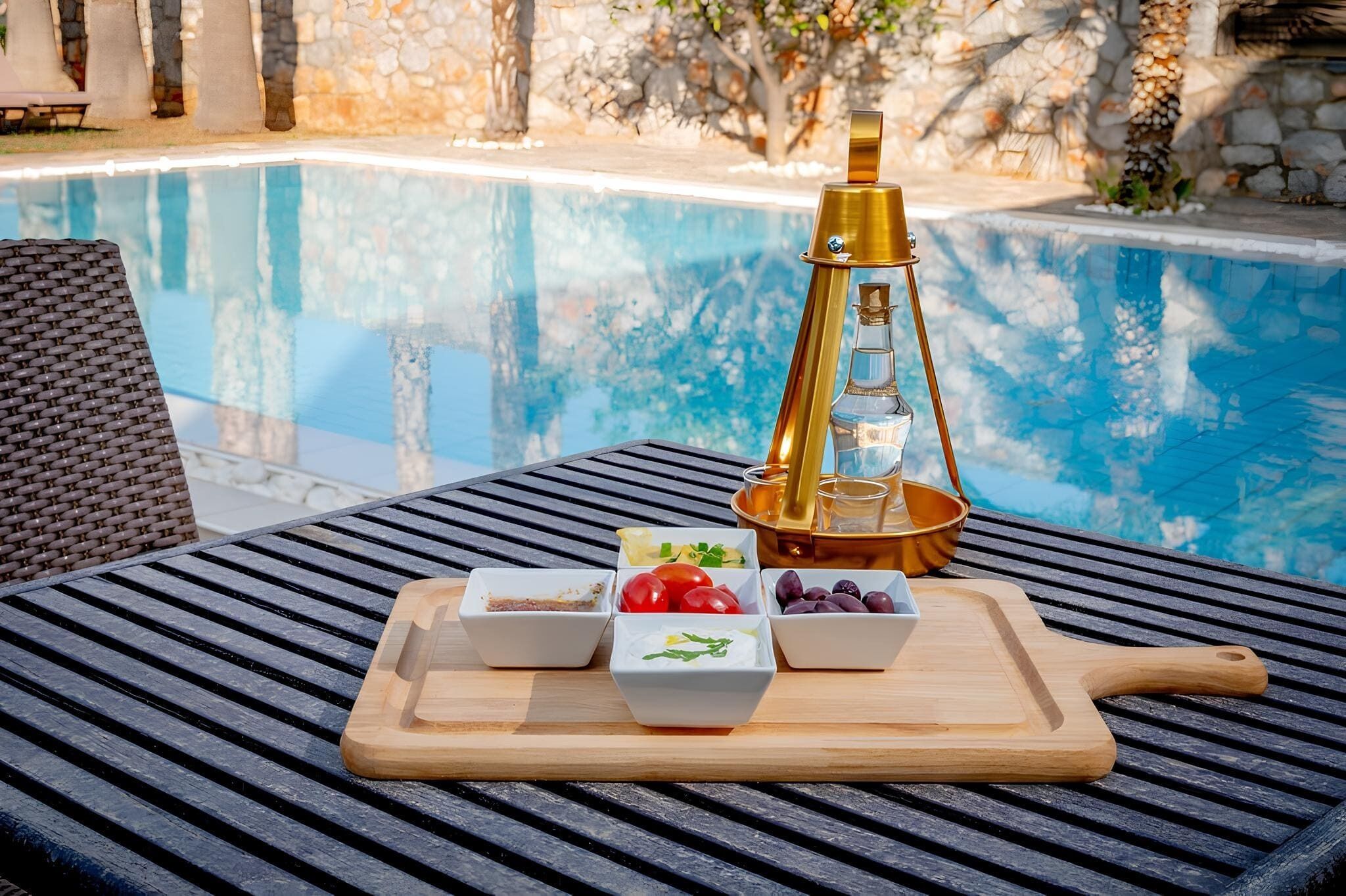 Mesa junto a la piscina con una tabla de madera que sostiene alimentos y una lámpara decorativa.