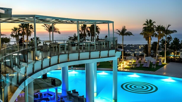 A modern pool area with a glass balustrade, lounge chairs, and a view of the ocean at sunset. - The Noverian Scenic Crete 5 Star Hilltop Villa Resort & Spa (Hersonissos)