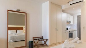 A hotel room with a bed, a mirror, a small wooden bench, a trash bin, and a kitchenette with cabinets and appliances. - The Noverian Scenic Crete 5 Star Hilltop Villa Resort & Spa (Hersonissos)