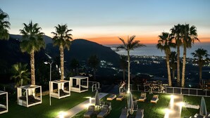 A resort with outdoor seating areas, palm trees, and a view of a city at sunset. - The Noverian Scenic Crete 5 Star Hilltop Villa Resort & Spa (Hersonissos)