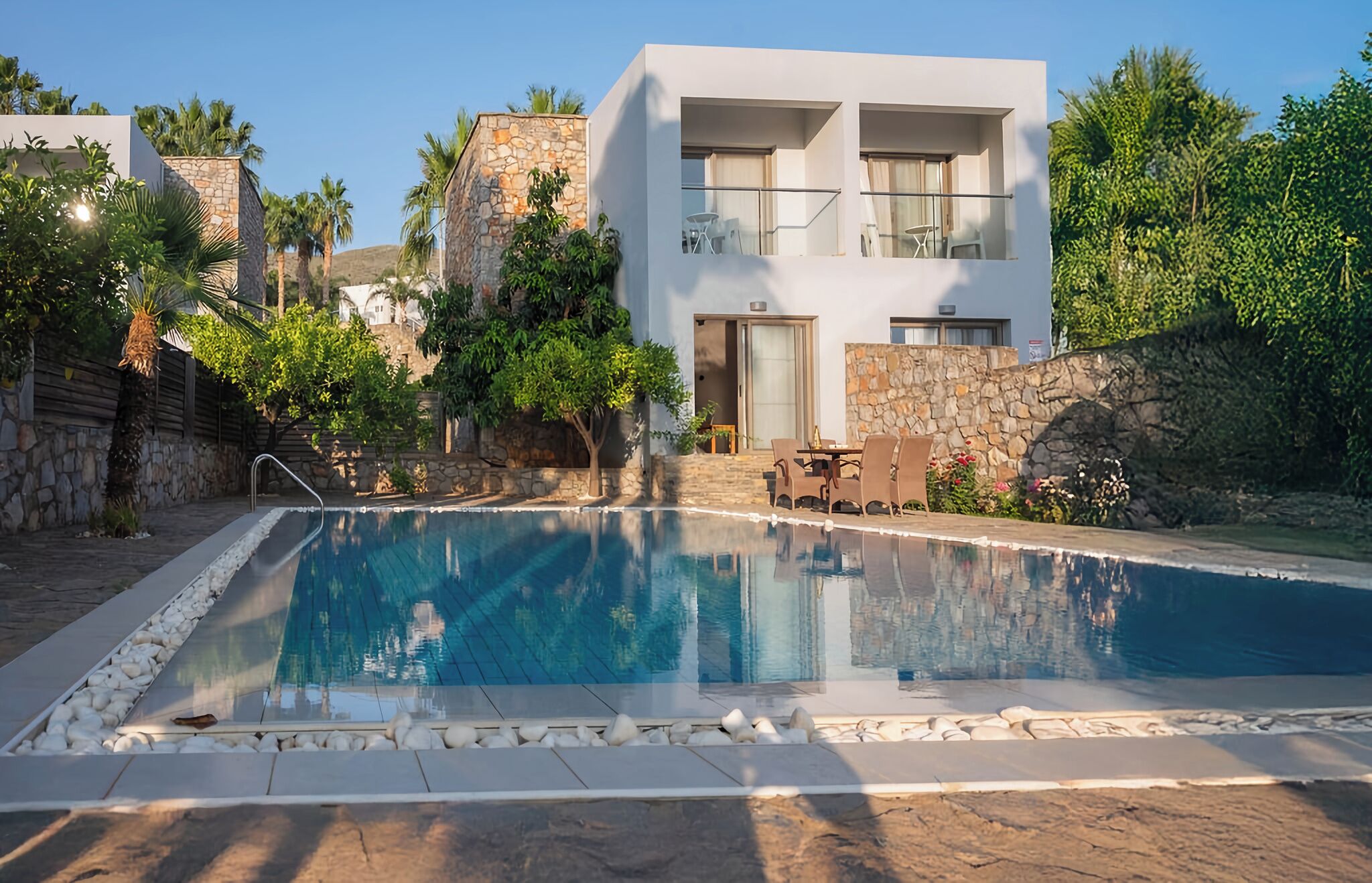 Un área de piscina moderna con una piscina azul cristalina, paredes de piedra y un edificio de dos pisos con balcones.