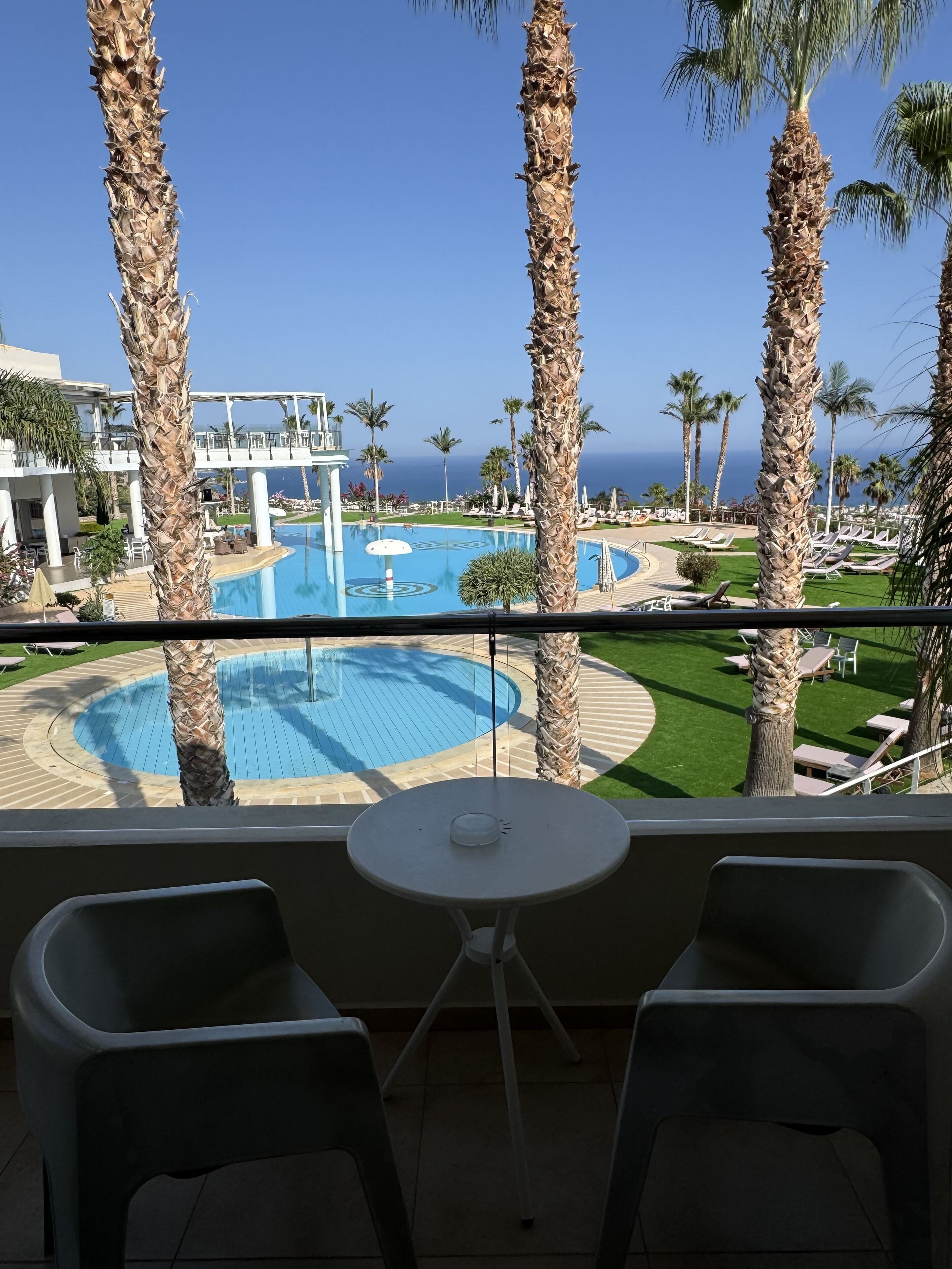 A balcony with two chairs and a small table, overlooking a pool area with palm trees and a view of the ocean.