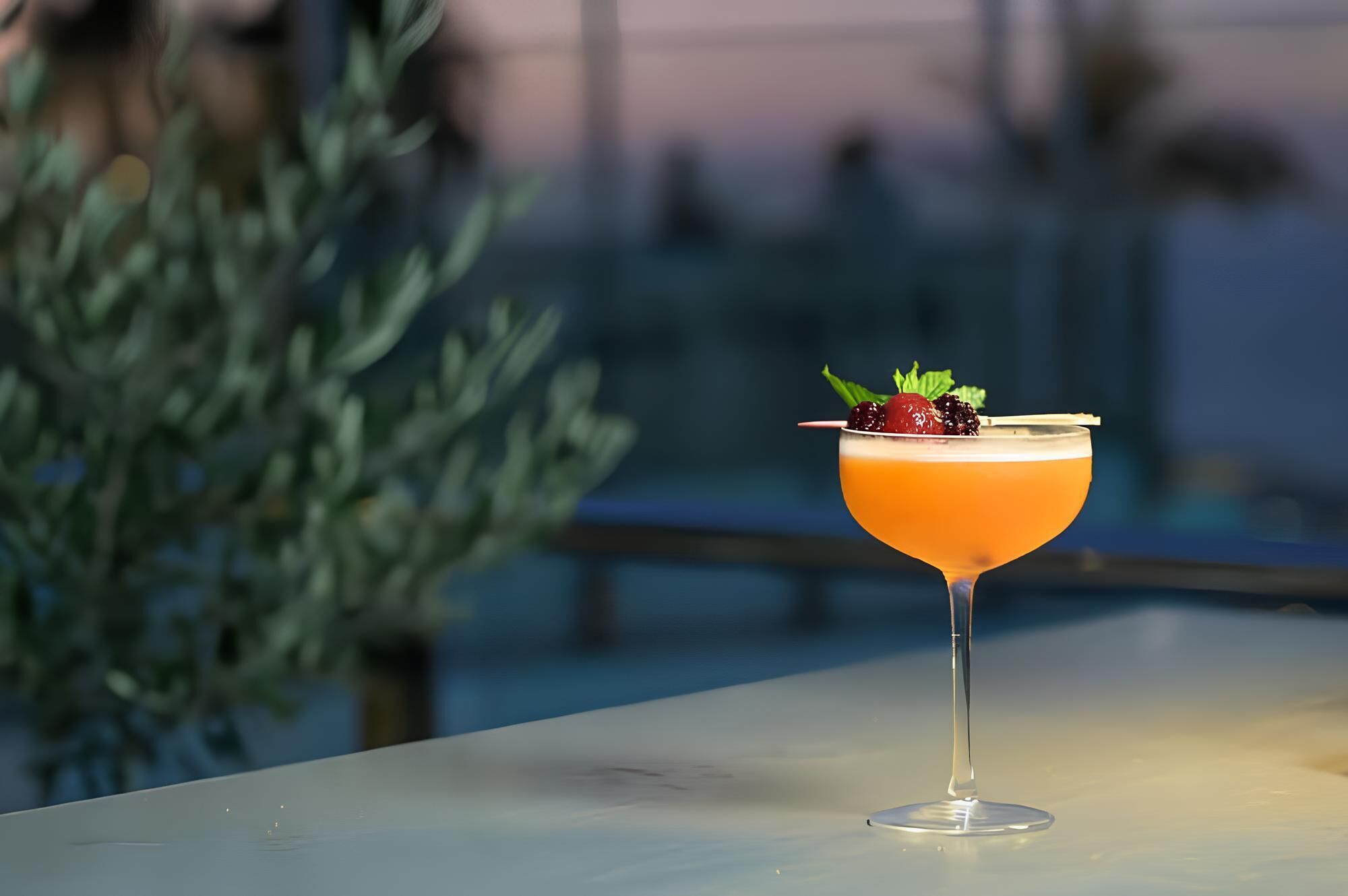 A cocktail in a stemmed glass with a garnish of berries and mint leaves, placed on a table with a blurred background.