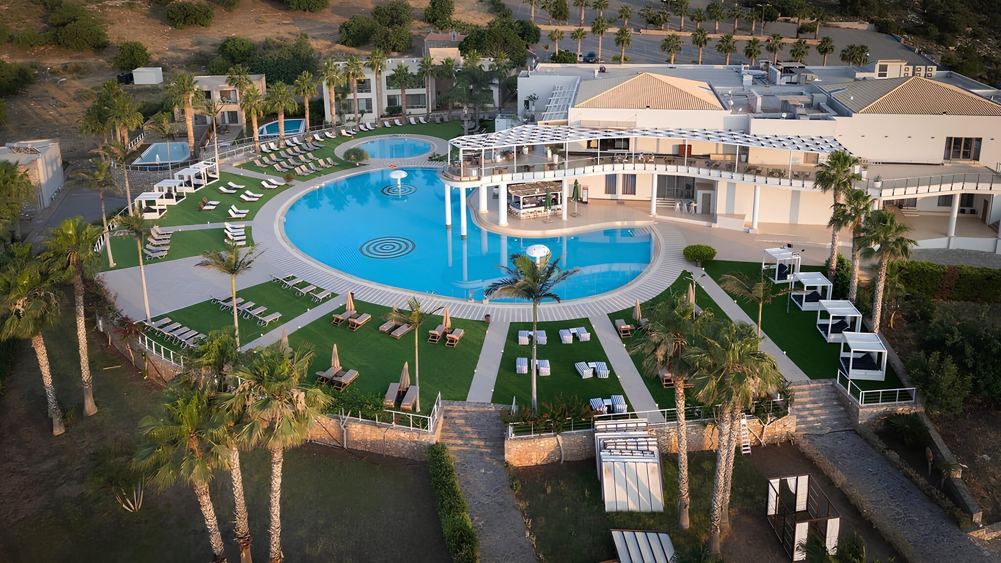 A resort with a large central pool, multiple lounge areas, and palm trees.