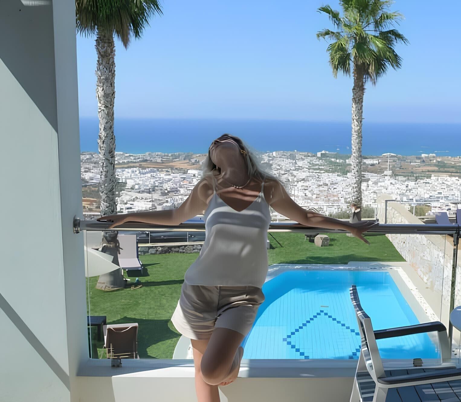 A person standing by a balcony with a pool and a view of a coastal town.