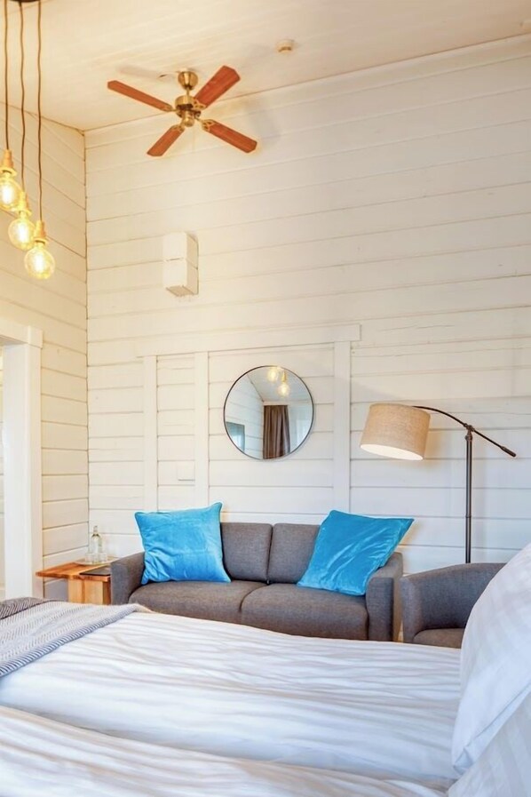 A bedroom with a bed, a gray sofa with blue cushions, a round mirror, a ceiling fan, and a floor lamp. - Hotel Breiðdalsvík (Fjardabyggd)