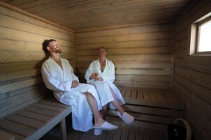 Duas pessoas em uma sauna de madeira com bancos e uma janela.