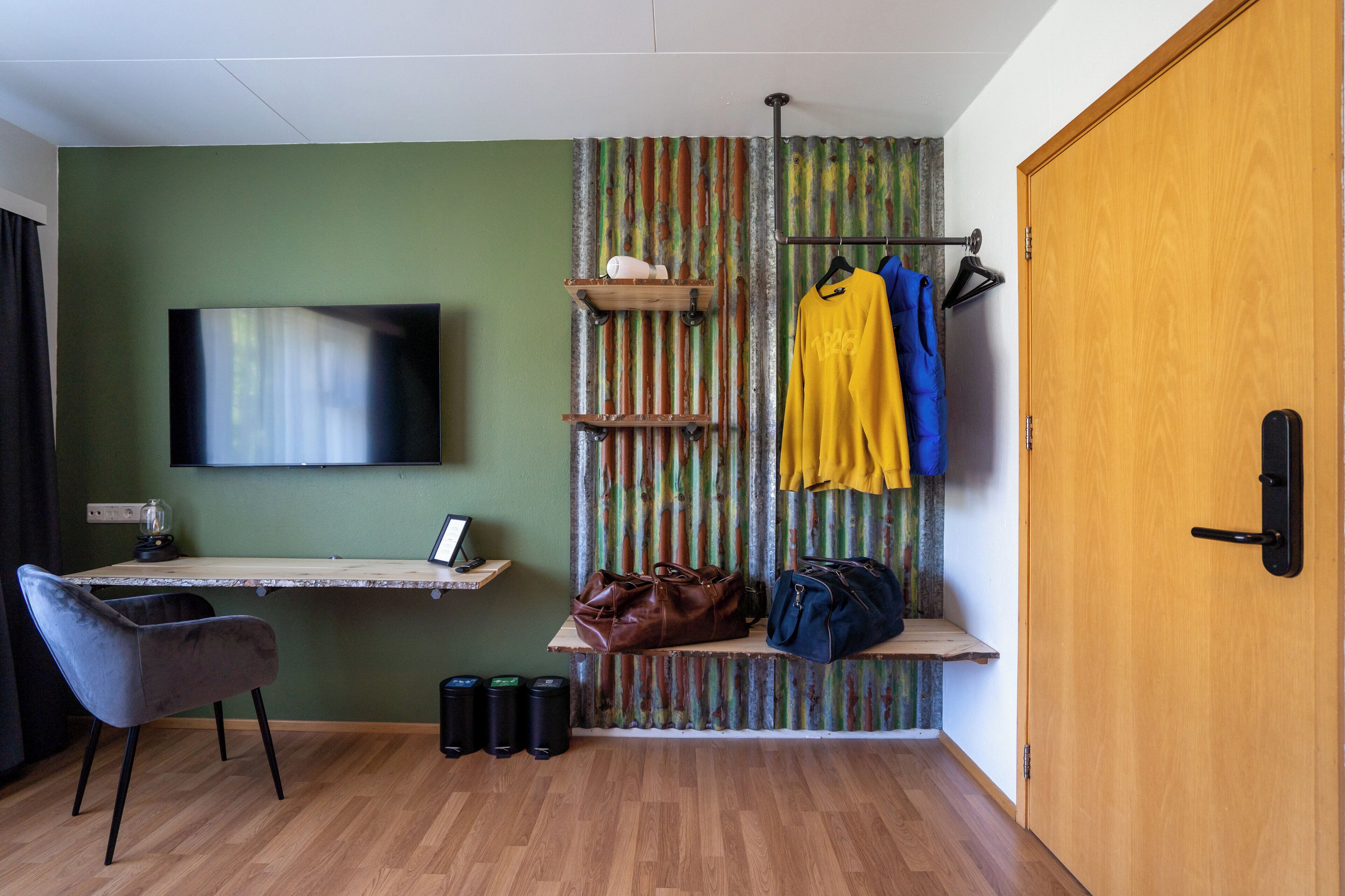 Una habitación con un escritorio de madera, una silla, un televisor montado en la pared, un perchero con chaquetas y una puerta.