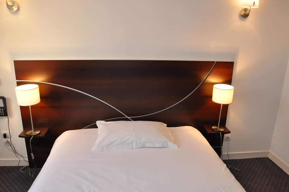 A hotel room with a large bed, two bedside lamps, a headboard with a curved design, and a plain wall.