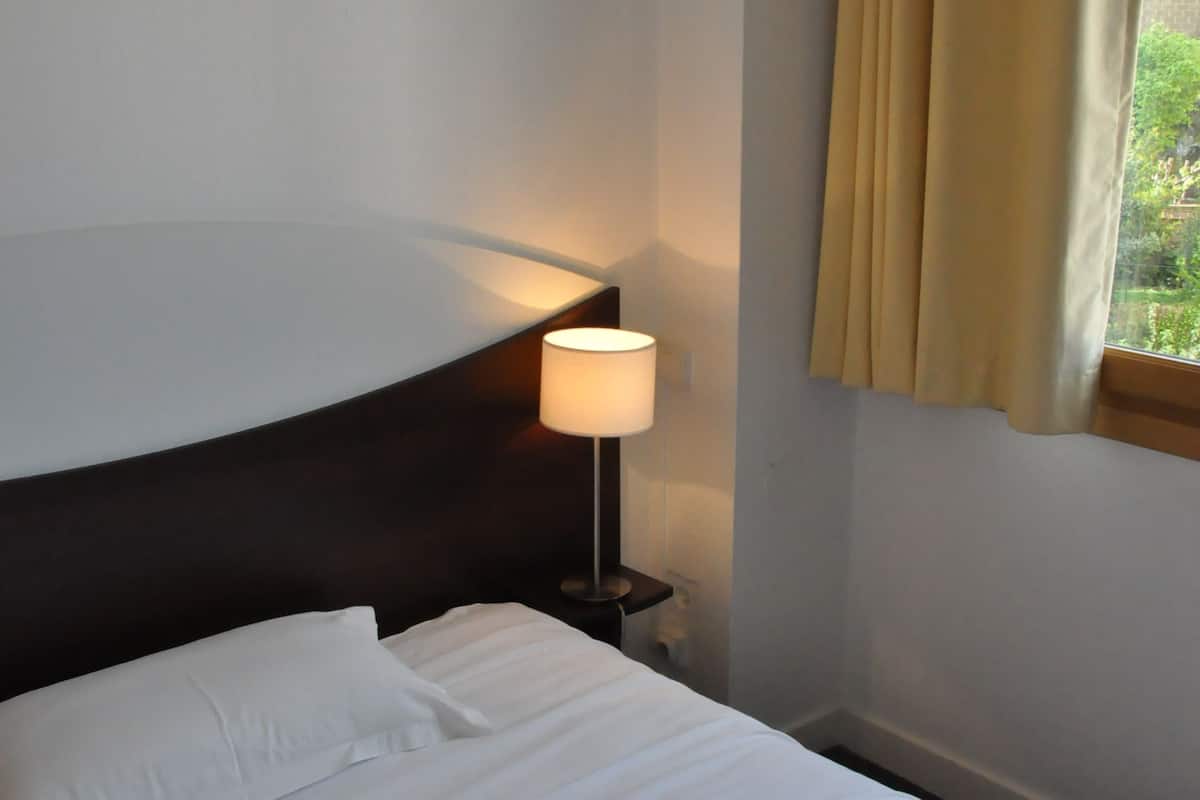 A neatly made bed with white linens, a bedside lamp, and a window with curtains.