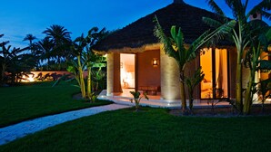 A thatched-roof building with a well-lit interior, surrounded by palm trees and a grassy area. - Les 5 Djellabas (Marrakech)