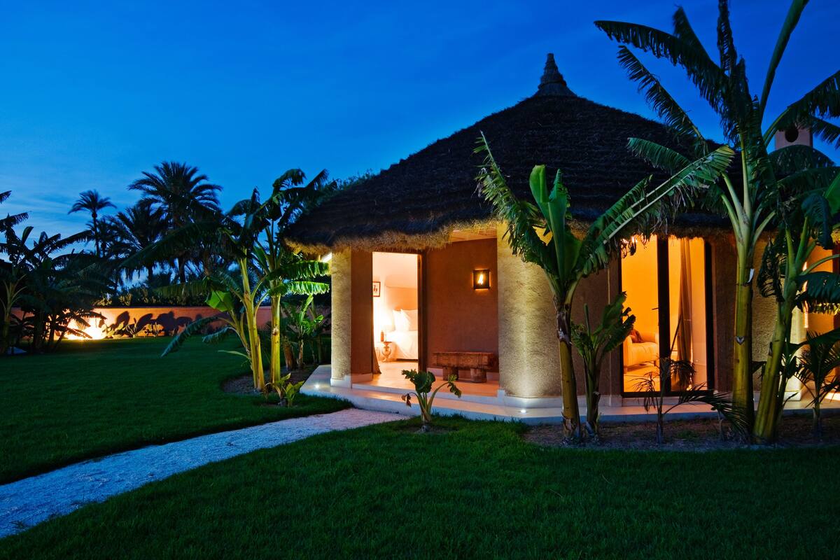 A thatched-roof building with a well-lit interior, surrounded by palm trees and a grassy area.