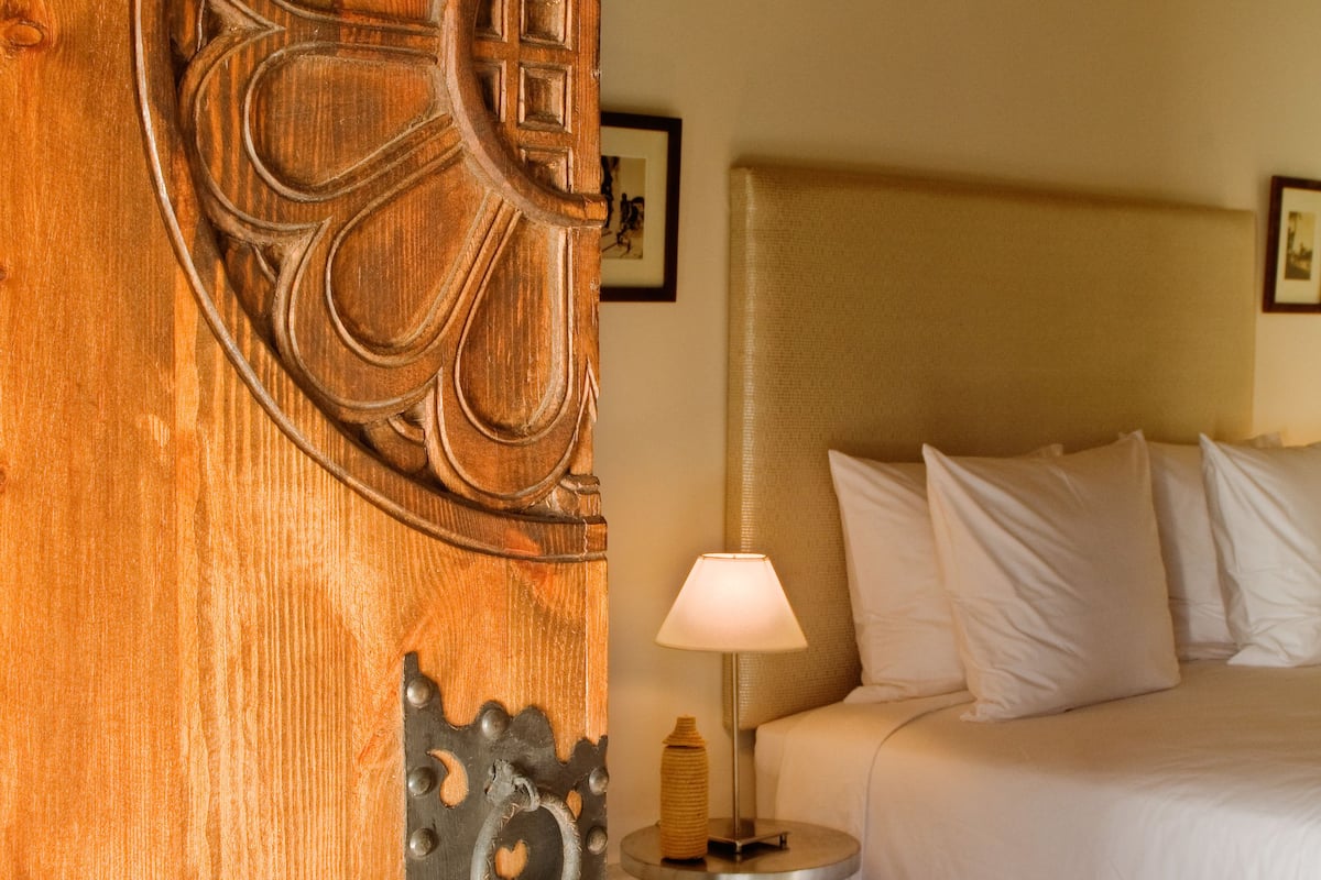 A wooden door with intricate carvings, a bed with white linens, a bedside lamp, and a headboard.