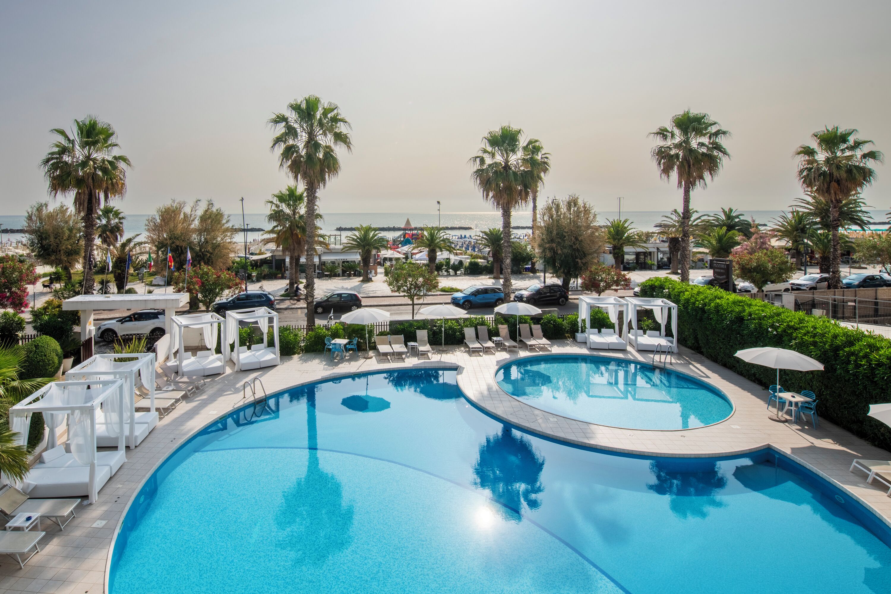 A large, curved swimming pool with lounge chairs and umbrellas, surrounded by palm trees and a view of the sea.