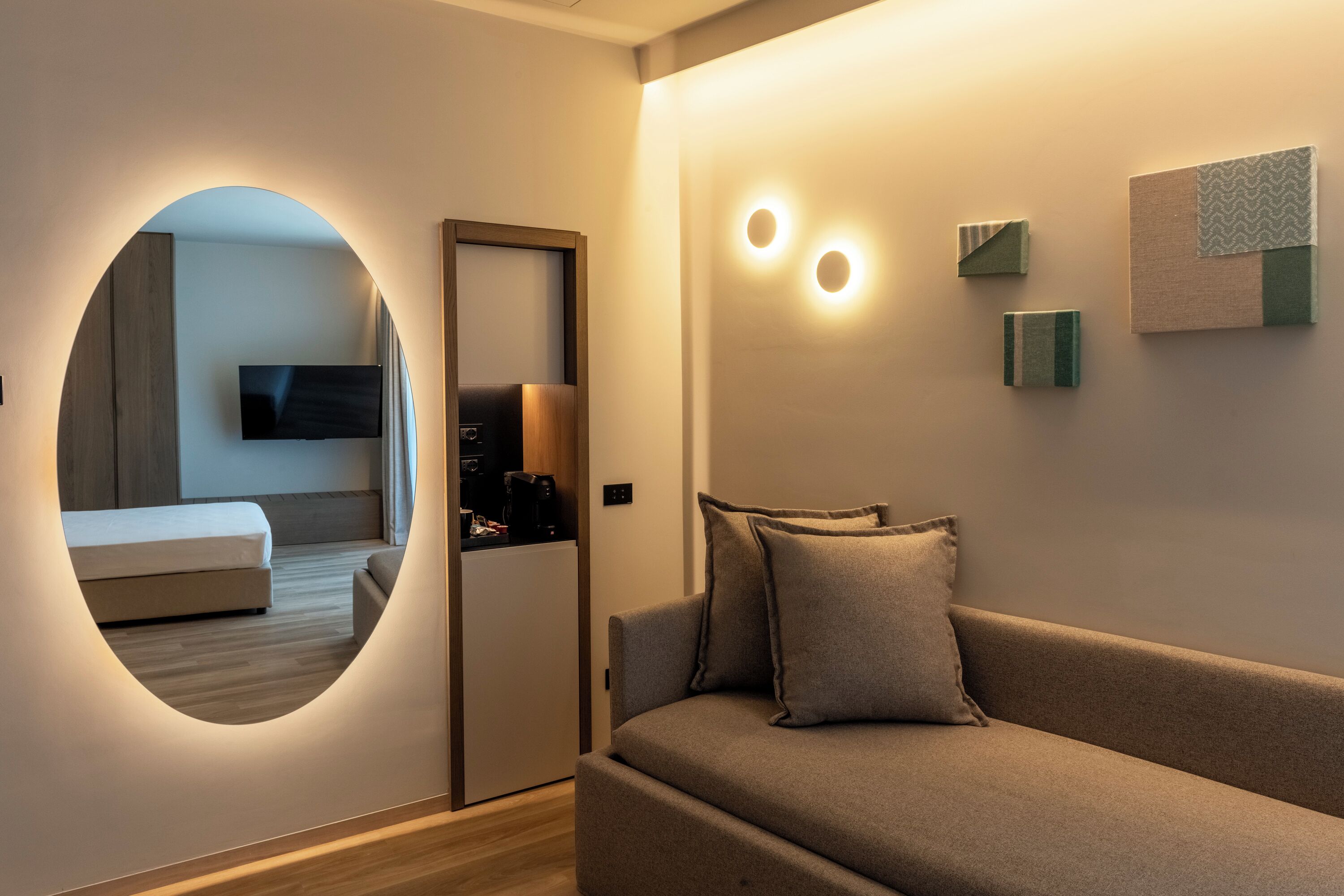 A modern hotel room with a circular mirror, a sofa, a flat-screen TV, and a small coffee machine.