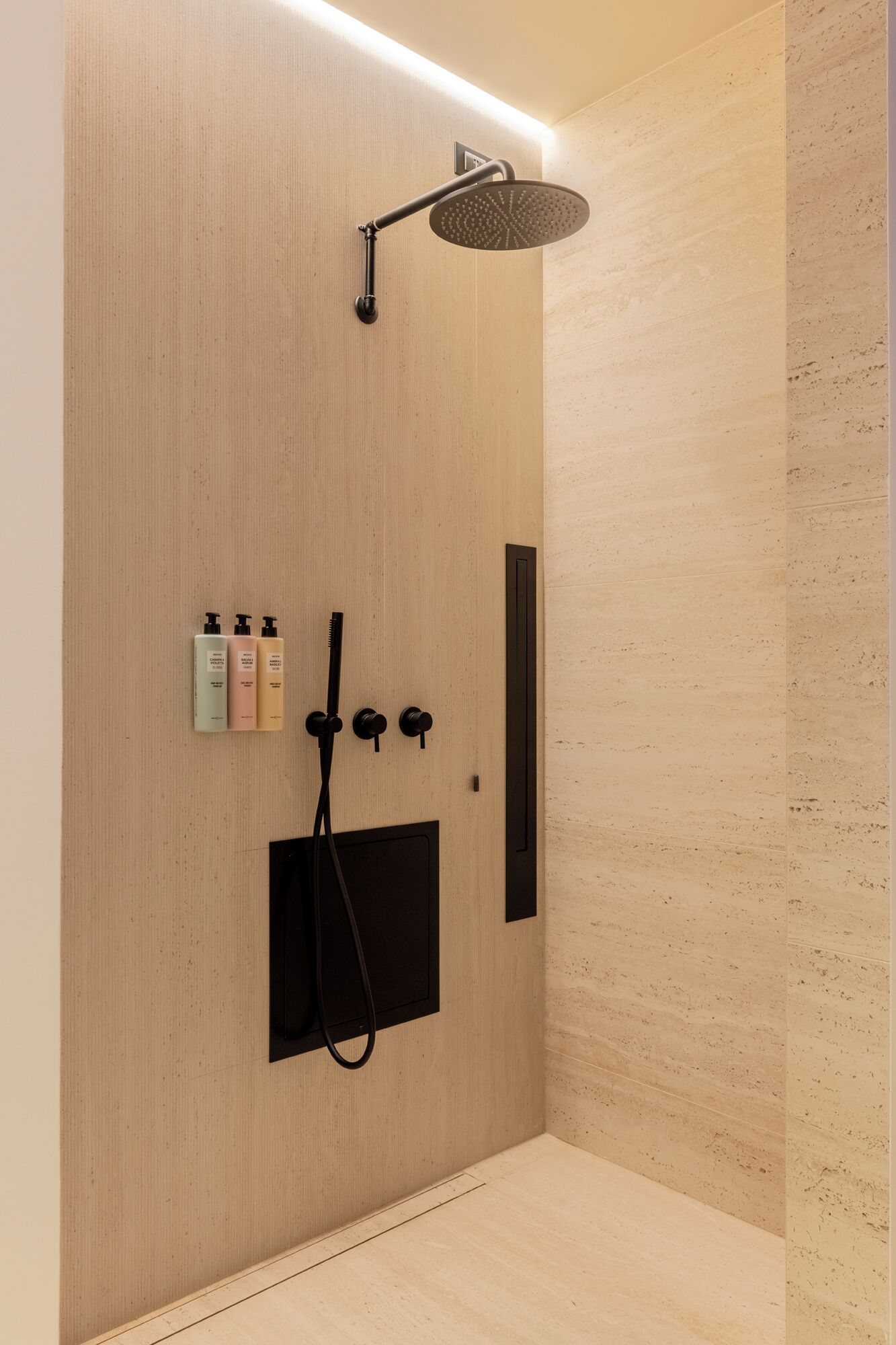 A modern shower with a rain showerhead, a handheld showerhead, and a control panel with three knobs. There are also three bottles of shampoo or conditioner on the wall.