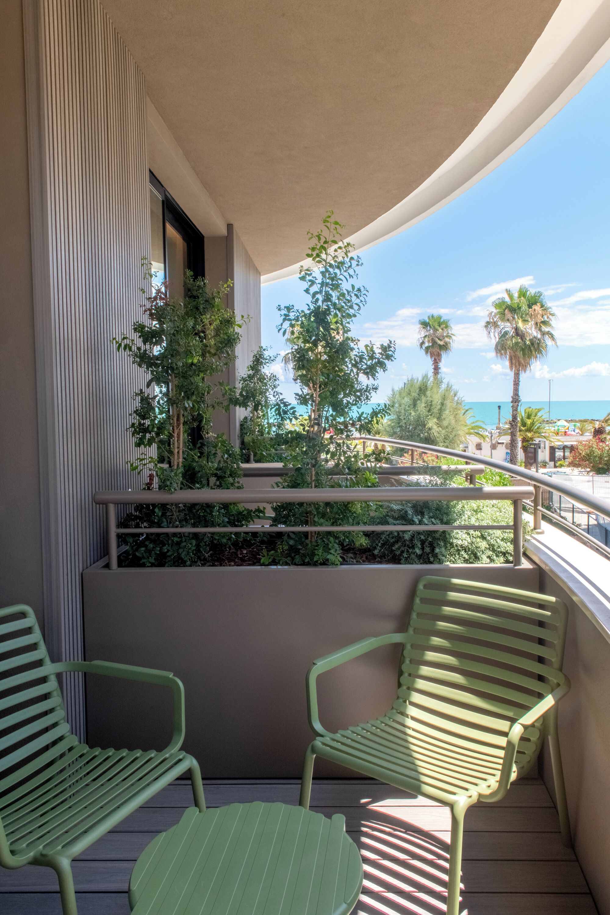 A balcony with two green chairs and a small table, overlooking a landscaped area with trees and a view of the beach.
