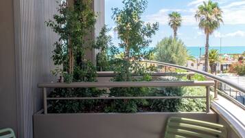 A balcony with two green chairs and a small table, overlooking a landscaped area with trees and a view of the beach.