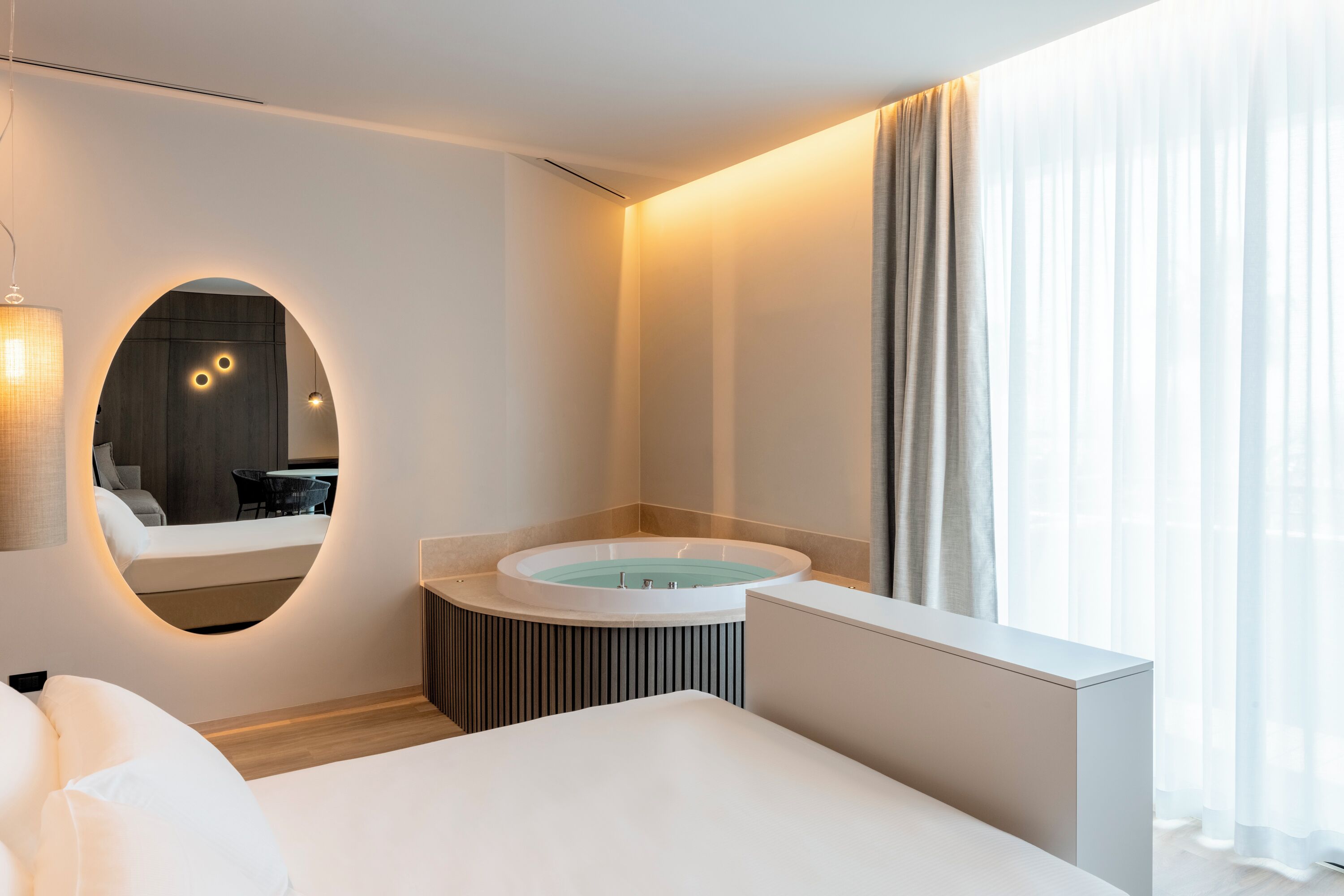 Habitación de hotel moderna con una cama grande, una bañera redonda, un espejo y vistas al exterior.