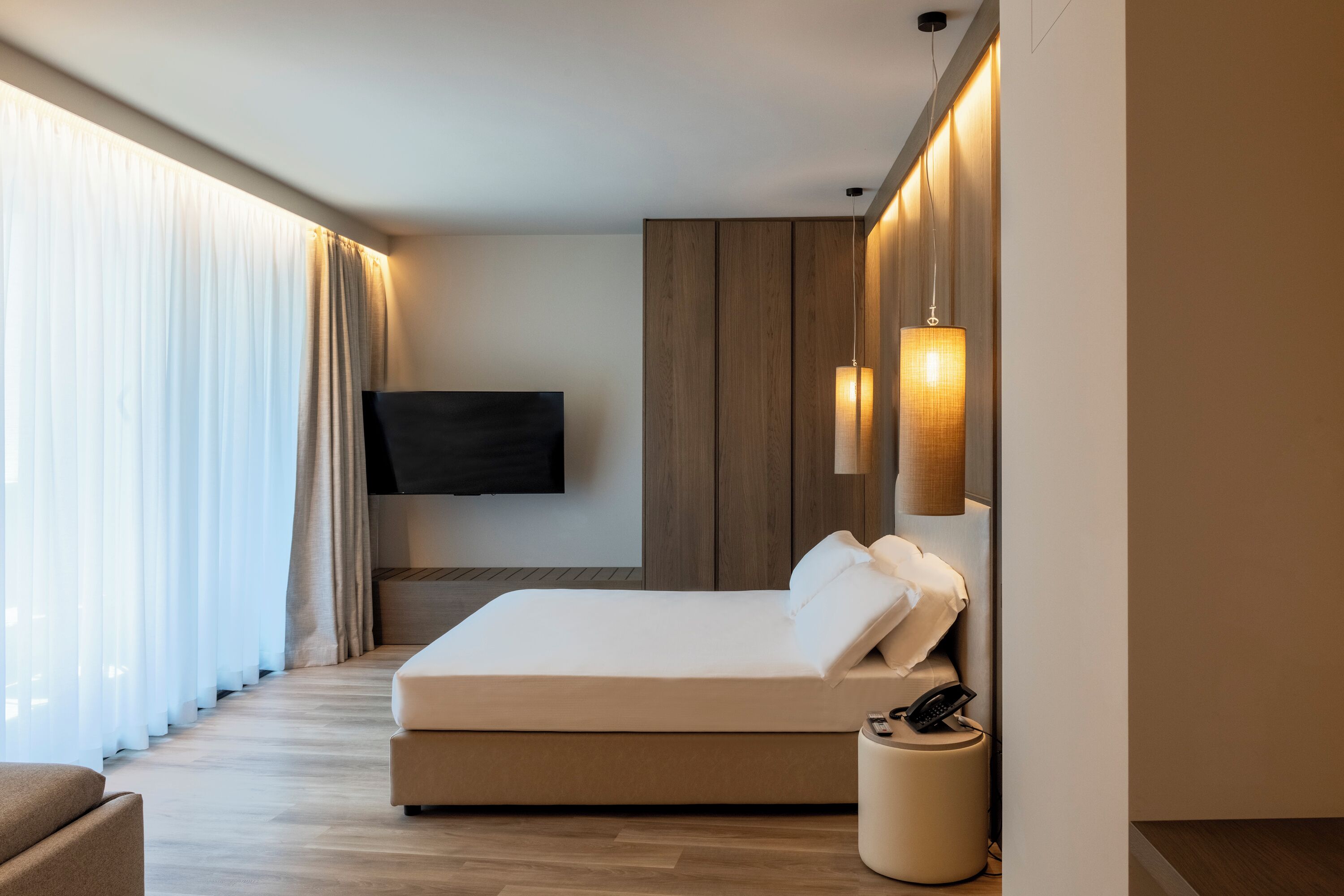 A modern hotel room with a large bed, a flat-screen TV mounted on the wall, and a bedside table with a lamp.
