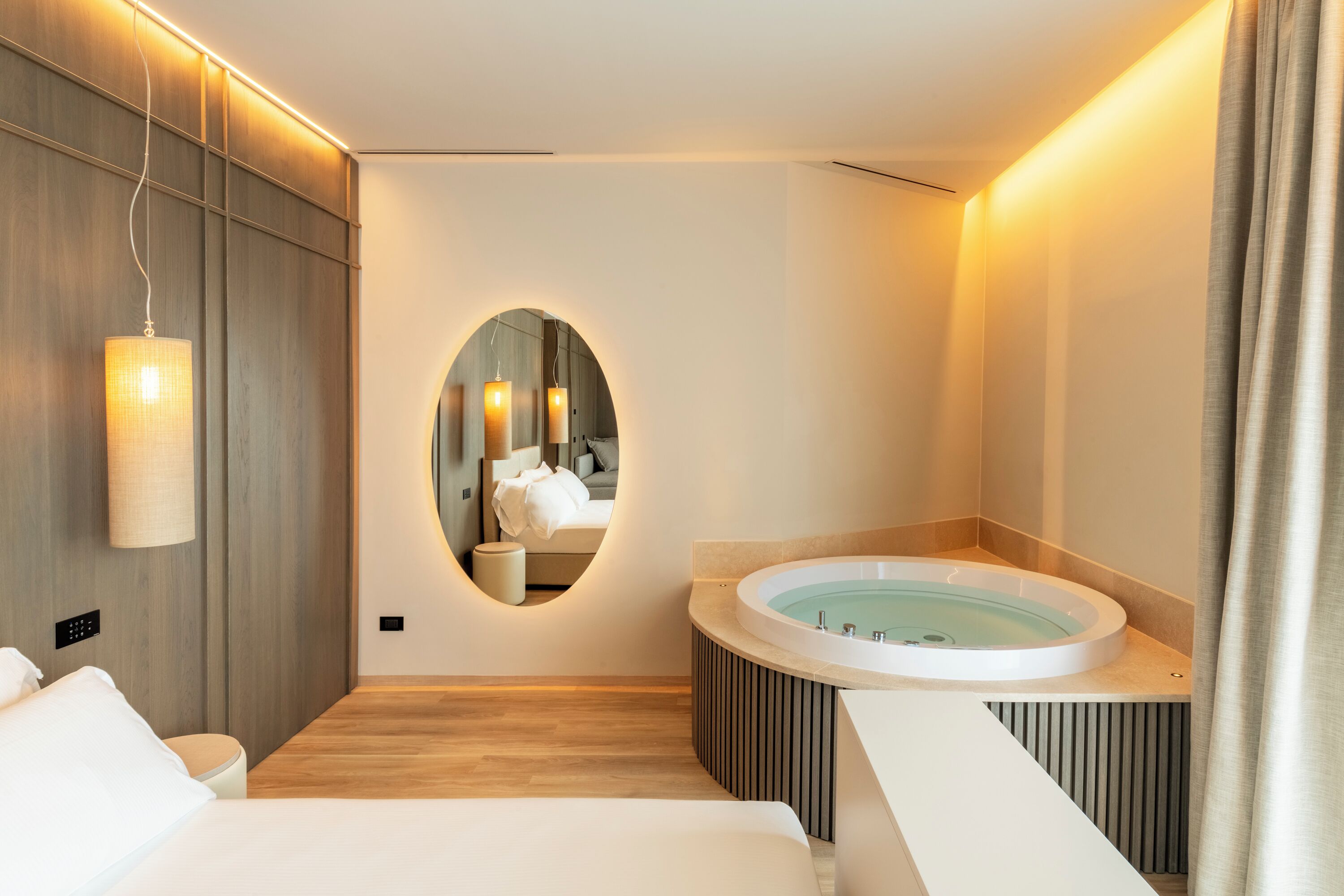A modern hotel room with a large bed, a round mirror, a built-in bathtub, and a small seating area.