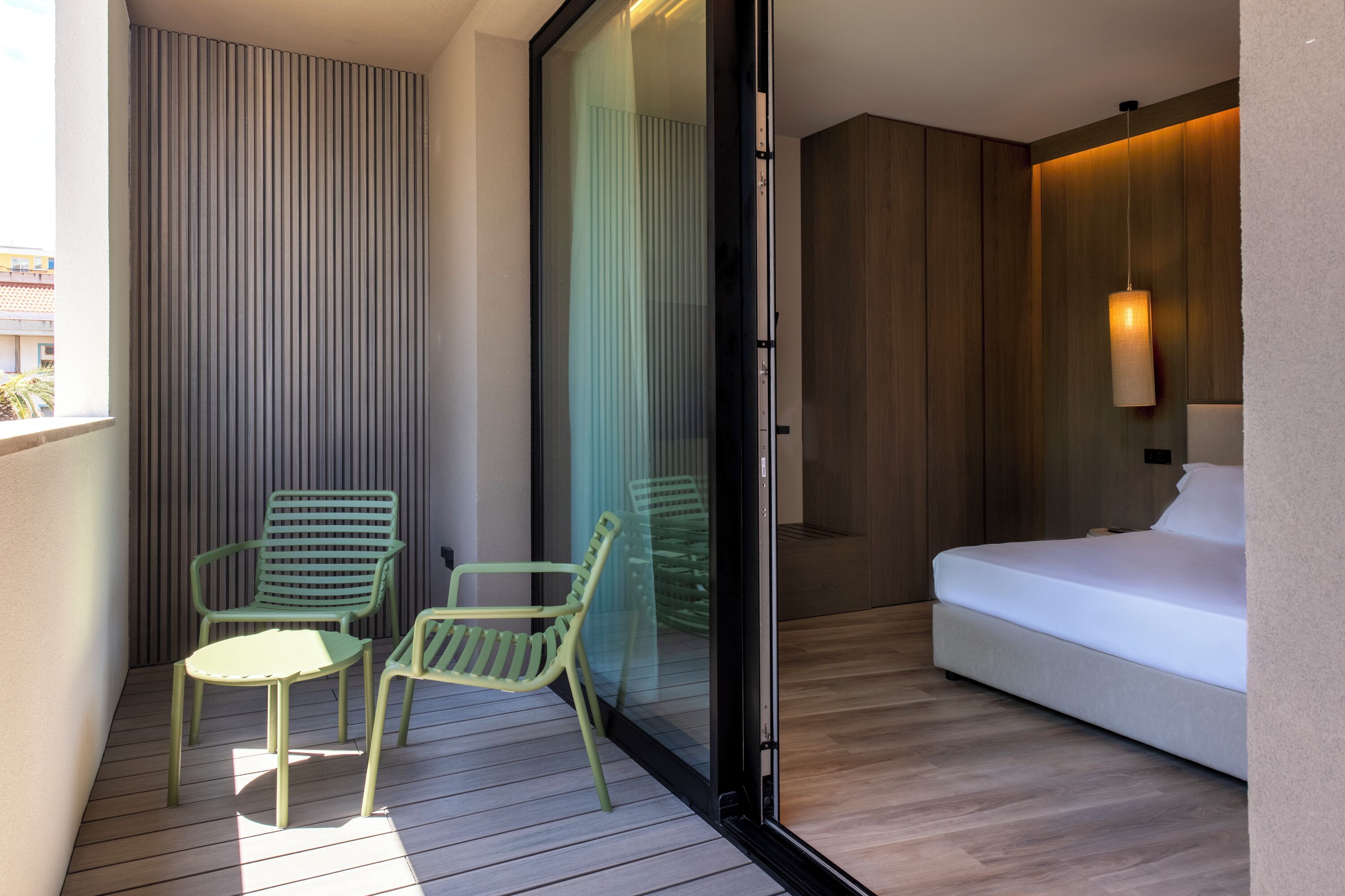 A modern hotel room with a sliding glass door leading to a balcony, a bed, and two chairs.